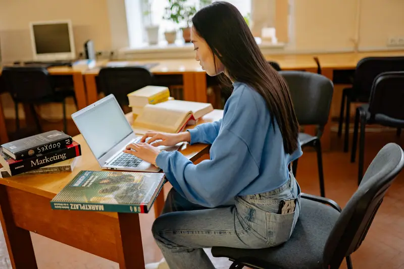 Voksen kvinne som studerer med bærbar PC og bøker ved et bord i et bibliotek